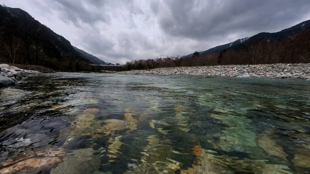 มุมมองระยะใกล้ของผิวน้ำในแม่น้ำอาซุสะ (Azusa River) คามิโคจิ ที่ใสจนมองเห็นก้อนหินก้นแม่น้ำได้อย่างชัดเจน ฉากหลังเป็นหุบเขาที่มืดครึ้มภายใต้ท้องฟ้ามีเมฆมาก