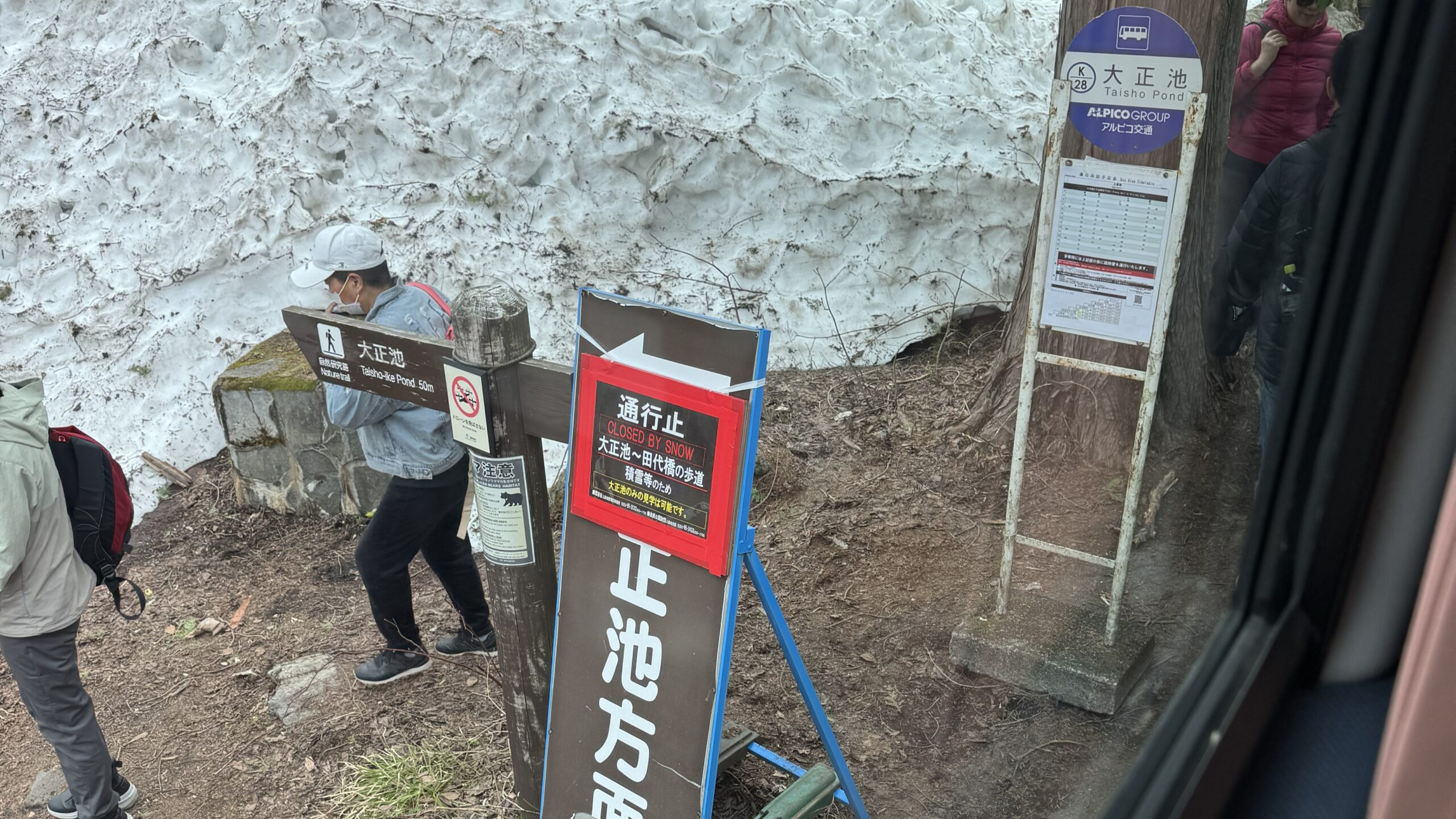 ภาพป้ายบอกทางและนักท่องเที่ยวที่จุดจอดรถบัส ป้าย Taisho Pond (大正池) ในคามิโคจิ (Kamikochi) โดยมีกำแพงหิมะสีขาวกองสูงอยู่ด้านหลัง บรรยากาศช่วงต้นฤดูใบไม้ผลิที่พื้นที่เพิ่งเปิดให้เข้าชม Caption (คำบรรยายใต้ภาพ) 🏔️ ช่วงเวลาที่หิมะยังไม่ละลายหมด: สัมผัสทิวทัศน์ฤดูใบไม้ผลิที่ไม่เหมือนใครที่จุดจอดรถในคามิโคจิ (Kamikochi