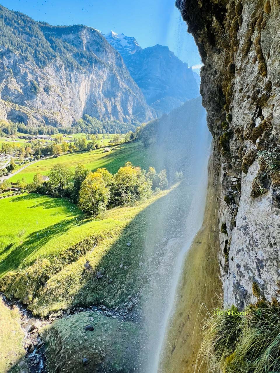 น้ำตกขนาดใหญ่ที่ไหลลงมาจากหน้าผาหินสูงลงสู่หุบเขาสีเขียวสดใสที่มีทุ่งหญ้าและต้นไม้ พร้อมฉากหลังเป็นภูเขาที่ปกคลุมด้วยหิมะเล็กน้อยภายใต้ท้องฟ้าสีครามสดใสในสวิตเซอร์แลนด์
