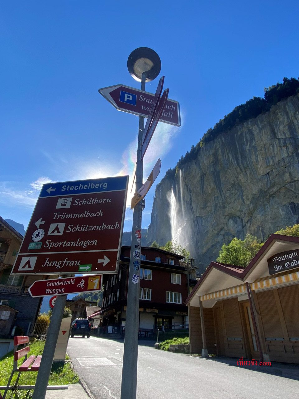 ป้ายบอกทางเดินป่าหลายทิศทางในหมู่บ้านสวิส โดยมีน้ำตกขนาดใหญ่และหน้าผาหินสูงชันเป็นฉากหลัง ภายใต้แสงแดดจ้าและท้องฟ้าสีคราม