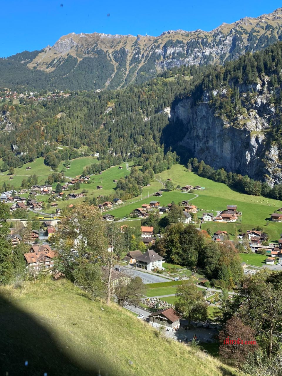 ทิวทัศน์หมู่บ้านในหุบเขา Lauterbrunnen ของสวิตเซอร์แลนด์ บ้านเรือนสไตล์สวิสตั้งอยู่บนทุ่งหญ้าเขียวขจี มีหน้าผาหินสูงชันและป่าสนทึบเป็นฉากหลัง ภายใต้ท้องฟ้าสีคราม