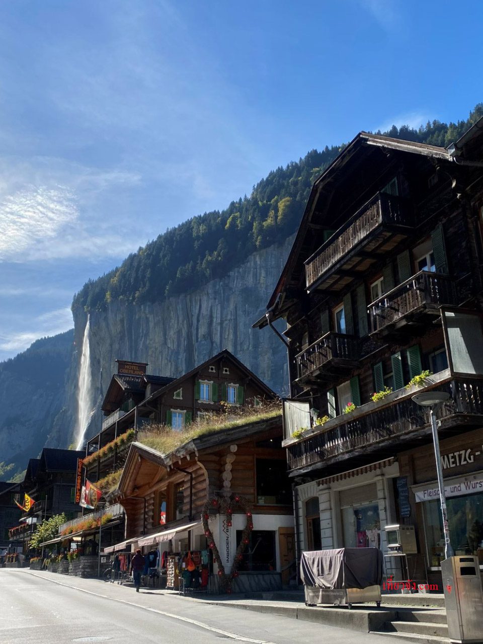 ถนนในหมู่บ้าน Lauterbrunnen สวิตเซอร์แลนด์ มีอาคารไม้สไตล์ Chalet แบบดั้งเดิมเรียงรายอยู่ด้านขวา ด้านซ้ายมีน้ำตกขนาดใหญ่พุ่งลงมาจากหน้าผาหินสูงชันภายใต้ท้องฟ้าสีคราม