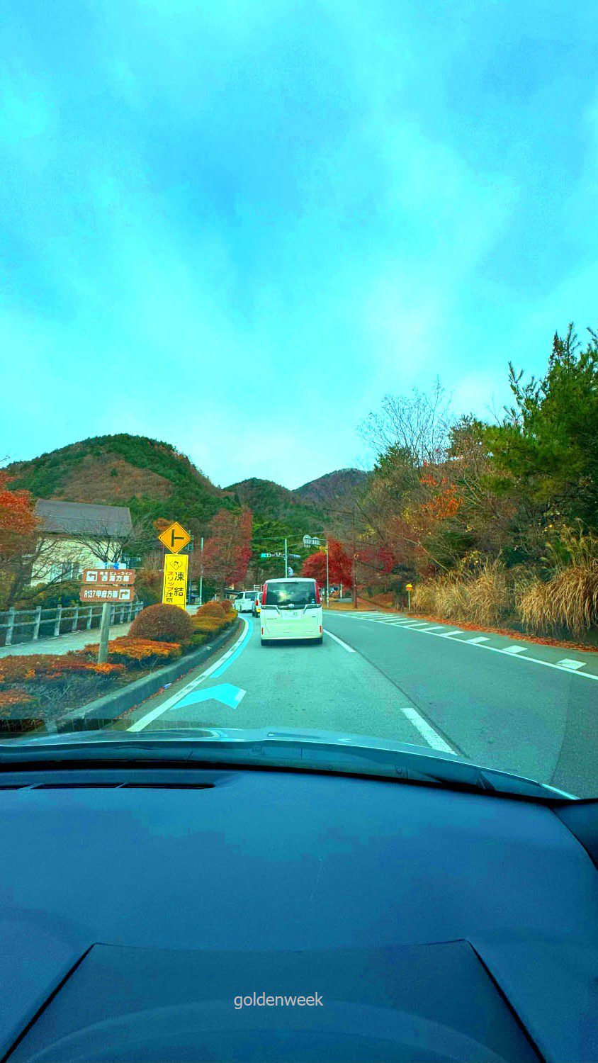 มุมมองจากรถยนต์บนถนนที่มีต้นไม้เปลี่ยนสีตามแนวภูเขาในฤดูใบไม้ร่วง ระหว่างเดินทางไป Kawaguchiko Momiji Kairo